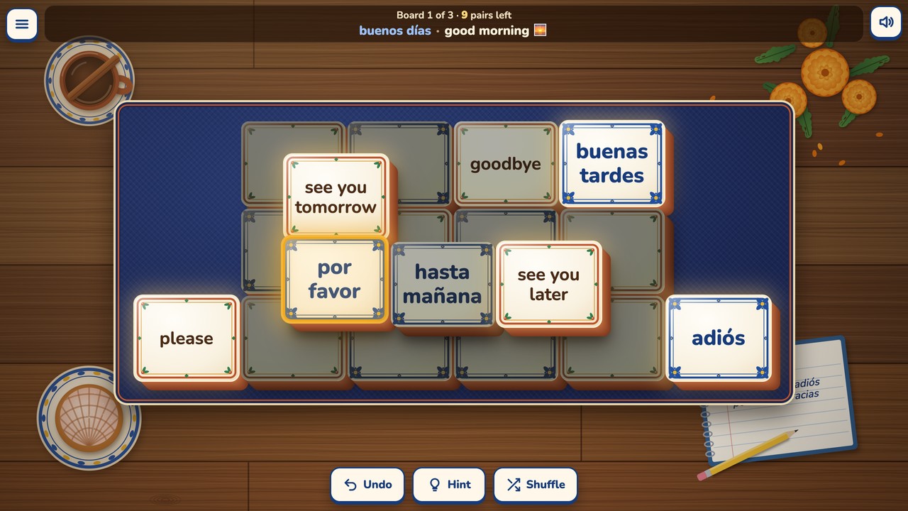 Board 1 part played on a blue woven mat on a café table. Spanish words sit on tiles with blue frames and English meanings on tiles with terracotta frames. Por favor is picked up with a gold rim, its partner please is bright and free at the left, the tiles in the shade (goodbye, hasta mañana) are blocked, and the status bar shows the last pair taken: buenos días · good morning, with a sunrise.